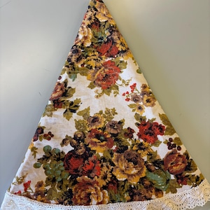 May include: Triangular floral tablecloth with a cream background and a pattern of red, yellow, and brown roses. The edge is trimmed with a white lace border. The tablecloth is folded.