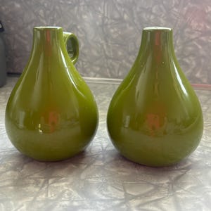 Może przedstawiać: Dwa zielone wazony ceramiczne z błyszczącym wykończeniem. Jeden wazon ma mały uchwyt. Wazony mają kształt łzy z wąską szyjką i szeroką podstawą. Wazony są prawdopodobnie do celów dekoracyjnych.