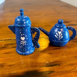 Puede incluir: Una tetera y una cafetera de cerámica azul y blanca con diseños florales. La tetera tiene un cuerpo redondo y un pico en el lateral. La cafetera tiene un cuerpo alto y cilíndrico y un pico en el lateral. Ambas piezas tienen un esmalte blanco moteado.