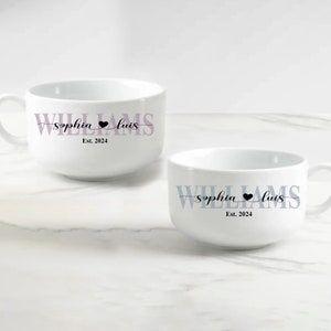 May include: Two white ceramic mugs featuring personalized text. One mug displays the name WILLIAMS in light purple with the script names Sophia and Luis separated by a black heart, and Est. 2024. The other mug features the same text in blue.