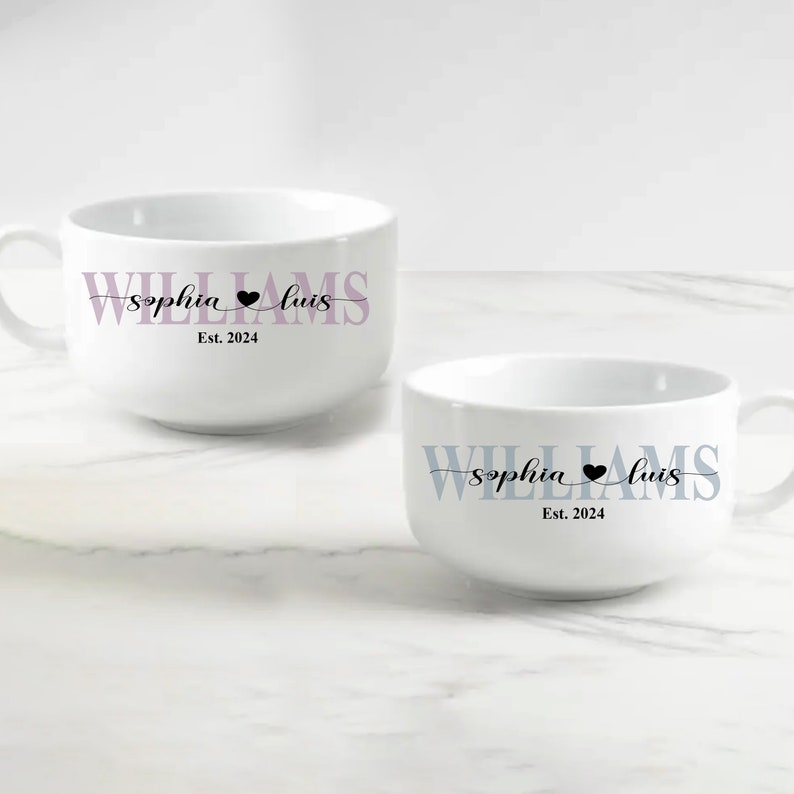 May include: Two white ceramic mugs featuring personalized text. One mug displays the name WILLIAMS in light purple with the script names Sophia and Luis separated by a black heart, and Est. 2024. The other mug features the same text in blue.