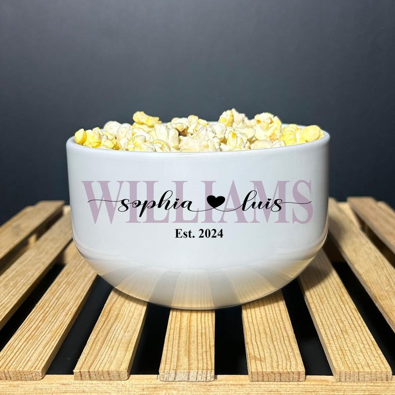 May include: A white ceramic bowl filled with popcorn. The bowl features the text "WILLIAMS" in large light purple letters, with the black script text "sophia &hearts; luis" and "Est. 2024" printed in the center of the bowl's exterior surface.