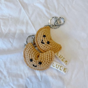 May include: Two crocheted fortune cookie keychains with smiling faces and the word "LUCK" embroidered on the white fabric attached to the key rings.
