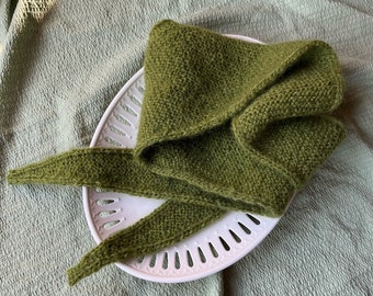 Può includere: Una sciarpa di maglia verde con estremità lunghe e fluttuanti, esposta su un piatto di ceramica bianca con tagli decorativi.