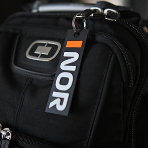 May include: Black travel backpack with a silver zipper and a rectangular tag that reads "NOR" in white letters. The tag has an orange accent at the top. The backpack has a silver logo on the front.