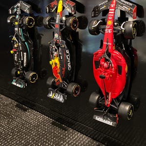 Op de afbeelding: Drie miniatuur Formule 1 raceauto's hangen aan een zwarte muur. De auto's zijn een zwarte Mercedes met Petronas branding, een rode Ferrari met Santander branding en een zwart-rode Red Bull met Oracle branding.