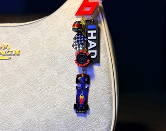 May include: A white handbag with a gold chain and a racing-themed charm. The charm includes a red number 6, a miniature helmet, a checkered flag, a tire, and a blue race car. The word "HAD" is printed on a black tag.