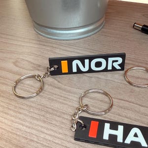 May include: Black keychains with silver key rings. The keychains have the word "INOR" in white letters with an orange rectangle. Another keychain has the word "HA" in white letters with a red rectangle.