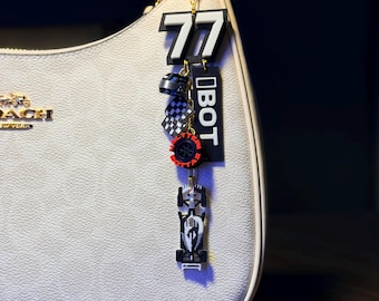 Può includere: Una borsa bianca con una catena dorata e un portachiavi a tema racing. Il portachiavi presenta il numero 77, la parola "BOT" e ciondoli in miniatura di auto e casco da corsa in nero, bianco e rosso.