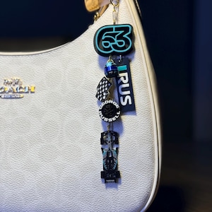 May include: A white handbag with a gold zipper and a charm. The charm features a teal and black number 63, a checkered flag, a wheel, a race car, and the word "RUS".
