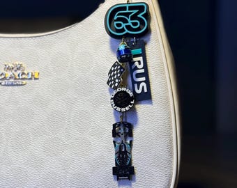 Könnte beinhalten: Eine weiße Handtasche mit goldenem Reißverschluss und einem Anhänger. Der Anhänger zeigt die teal-schwarze Zahl 63, eine Zielflagge, ein Rad, ein Rennwagen und das Wort "RUS".