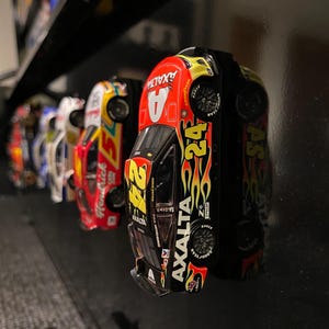 Op de afbeelding: Een verzameling van vijf miniatuur raceauto's die op een plank worden tentoongesteld. De auto's zijn in verschillende kleuren geverfd, waaronder rood, geel, blauw en zwart. De voorste auto is zwart met gele vlammen en de tekst "Axalta" aan de zijkant. De auto heeft het nummer "24" aan de zijkant.