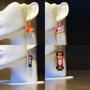 May include: Four pairs of earrings displayed on a white mannequin head. The earrings feature racing-themed designs: a red and yellow helmet, the number 16, the word "ILEC", and a race car. Each earring hangs from a silver hook.