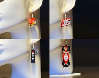 May include: Four pairs of earrings displayed on a white mannequin head. The earrings feature racing-themed designs: a red and yellow helmet, the number 16, the word "ILEC", and a race car. Each earring hangs from a silver hook.