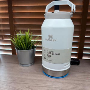 K&ouml;nnte beinhalten: Eine wei&szlig;e Stanley Flip Straw-Kanne mit grauem Deckel und Griff. Die Kanne hat einen silbernen Boden und einen blauen Ring. Der Text "STANLEY" und "THE ICEFLOW FLIP STRAW JUG" sind sichtbar. Links ist eine kleine Topfpflanze.