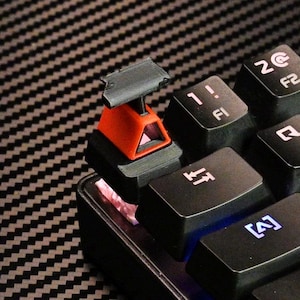 May include: A custom keycap shaped like a miniature oil derrick, in black and orange, is placed on a black keyboard. The keycap features a small platform and a stylized derrick structure. The keyboard keys are black with white lettering.