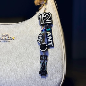 Op de afbeelding: Een witte handtas met een gouden ketting en een bedel. De bedel heeft het zwarte nummer 12, een geblokte vlag, een band, een raceauto en het woord "ANT". De handtas heeft een herhalend patroon.