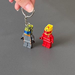 May include: Two Lego minifigure keychains. One gray and orange minifigure with a yellow helmet and blue face covering is attached to a silver keyring. The other is a red minifigure with a yellow helmet.