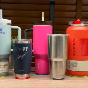 K&ouml;nnte beinhalten: Eine Sammlung isolierter Becher in verschiedenen Farben, darunter Hellblau, Marineblau, Pink, Edelstahl und Orange. Die Becher haben unterschiedliche Designs, einige mit den Logos von Stanley und Yeti. Einige Becher haben Griffe und Strohhalme.