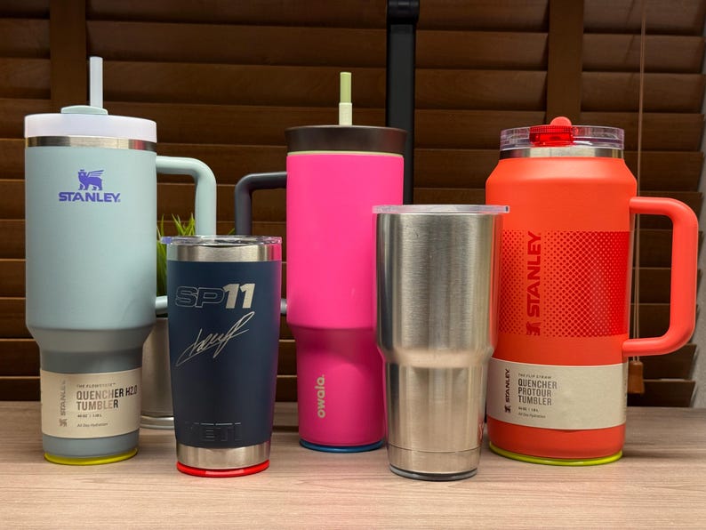 K&ouml;nnte beinhalten: Eine Sammlung isolierter Becher in verschiedenen Farben, darunter Hellblau, Marineblau, Pink, Edelstahl und Orange. Die Becher haben unterschiedliche Designs, einige mit den Logos von Stanley und Yeti. Einige Becher haben Griffe und Strohhalme.