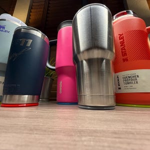 K&ouml;nnte beinhalten: Eine Auswahl an isolierten Bechern und Wasserflaschen in verschiedenen Farben, darunter Hellblau, Marineblau, Pink und Edelstahl. Die Marken sind Stanley, Yeti und Owala. Einige haben Griffe und Deckel. Auf der Stanley-Flasche steht "QUENCHER PROTOUR TUMBLER".