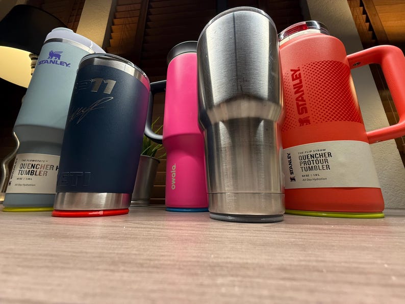 K&ouml;nnte beinhalten: Eine Auswahl an isolierten Bechern und Wasserflaschen in verschiedenen Farben, darunter Hellblau, Marineblau, Pink und Edelstahl. Die Marken sind Stanley, Yeti und Owala. Einige haben Griffe und Deckel. Auf der Stanley-Flasche steht "QUENCHER PROTOUR TUMBLER".