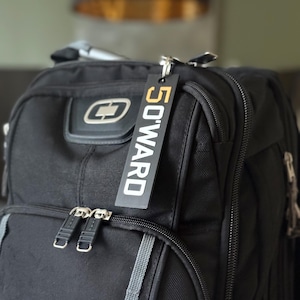 May include: Black backpack with silver zippers and a rectangular tag that reads "50WARD" in white letters on a black and gold background. The tag is attached to the backpack with a silver ring.