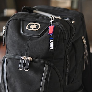 May include: Black backpack with silver zippers and a small red and blue keychain that says "VER". The backpack has a large front pocket and a smaller top pocket. The keychain is attached to the top zipper.