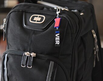 May include: Black backpack with silver zippers and a small red and blue keychain that says "VER". The backpack has a large front pocket and a smaller top pocket. The keychain is attached to the top zipper.