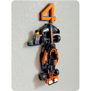 May include: A black and orange race car model with the number 4 in orange above it. A small Lego figure and a small Lego cube are on a black shelf attached to the car. The car has black wheels and orange accents.