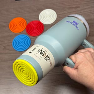 May include: A light blue Stanley Quencher H2.0 tumbler with a handle, holding a person's hand. The tumbler has a yellow base and a white lid. There are five colorful silicone coasters: orange, red, blue, white. The tumbler is 40 oz.