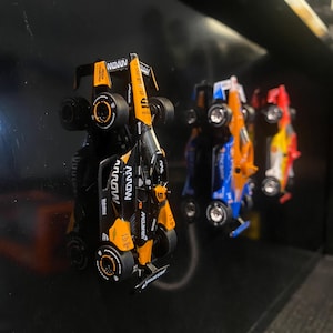 Op de afbeelding: Een verzameling van vier miniatuur raceauto's, waaronder een zwarte en oranje McLaren Formule 1-auto met het woord "NOVON" aan de zijkant, een blauwe en oranje auto, een rode en gele auto en een gele en zwarte auto. De auto's worden getoond op een zwart oppervlak.
