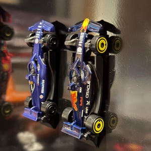 Op de afbeelding: Twee miniatuur Formule 1 raceauto's, een blauwe en een rode, worden getoond op een zwart oppervlak. De blauwe auto heeft de tekst "HONDA" en "YOKOHAMA" aan de zijkant. De rode auto heeft de tekst "Red Bull" en "ORACLE" aan de zijkant.
