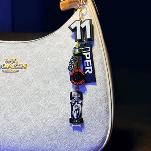 Könnte beinhalten: Eine weiße Handtasche mit goldener Kette und einem Anhänger. Der Anhänger zeigt die Zahl 11, ein schwarz-rotes Design und das Wort "PEREZ". Die Tasche hat ein sich wiederholendes Muster und ein goldenes Coach-Logo.