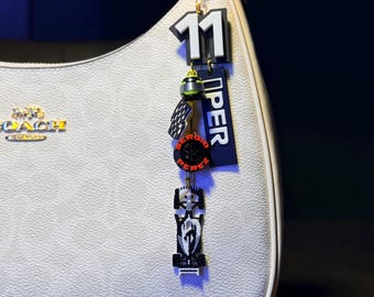 May include: A white handbag with a gold-tone chain and a charm. The charm features the number 11, a black and red design, and the word "PEREZ". The bag has a repeating pattern and a gold Coach logo.