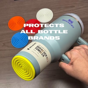 K&ouml;nnte beinhalten: Ein hellblauer Stanley Quencher H2.0 Becher mit Griff, gehalten von einer Hand. Der Becher hat einen gelben Boden und einen wei&szlig;en Deckel. Vier Untersetzer aus Silikon in Orange, Rot, Blau und Wei&szlig; befinden sich neben dem Becher. Der Text auf dem Bild lautet: PROTECTS ALL BOTTLE BRANDS.
