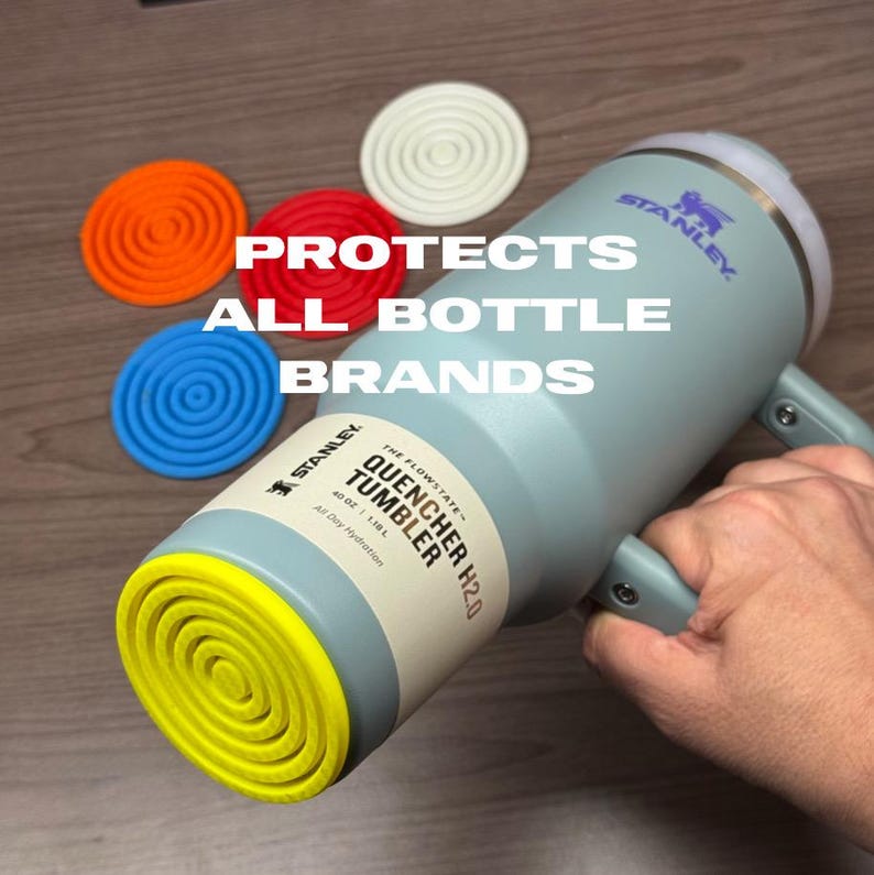 K&ouml;nnte beinhalten: Ein hellblauer Stanley Quencher H2.0 Becher mit Griff, gehalten von einer Hand. Der Becher hat einen gelben Boden und einen wei&szlig;en Deckel. Vier Untersetzer aus Silikon in Orange, Rot, Blau und Wei&szlig; befinden sich neben dem Becher. Der Text auf dem Bild lautet: PROTECTS ALL BOTTLE BRANDS.