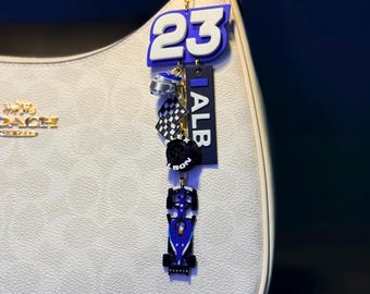May include: A keychain with a racing theme, featuring a blue and white number 23, a checkered flag, and a miniature race car. The keychain also includes a tag that says "ALB". The keychain is attached to a white handbag with gold hardware.