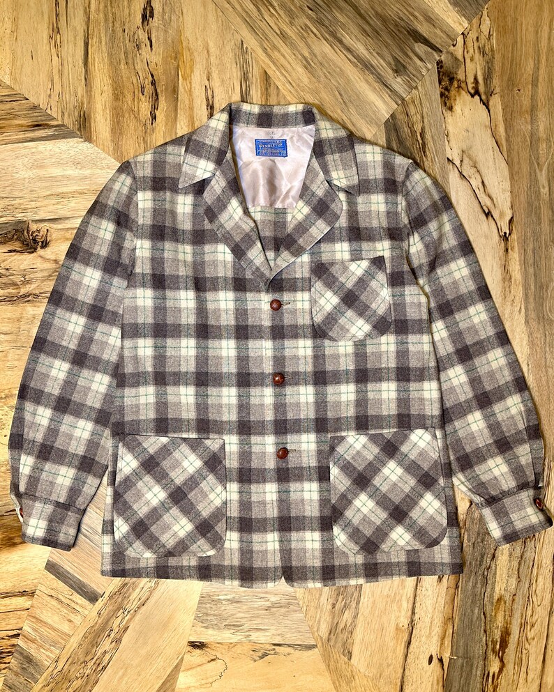 Puede incluir: Chaqueta vintage a cuadros en tonos grises, blancos y marrones. La chaqueta presenta un cuello cl&aacute;sico, dos bolsillos delanteros y botones marrones. La tela tiene una textura tejida.