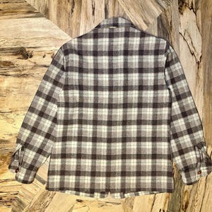 Puede incluir: Una camisa de manga larga con botones, con un dise&ntilde;o de cuadros en tonos marr&oacute;n, gris y crema. La camisa tiene cuello y pu&ntilde;os abotonados. La tela parece ser un material suave y tejido.