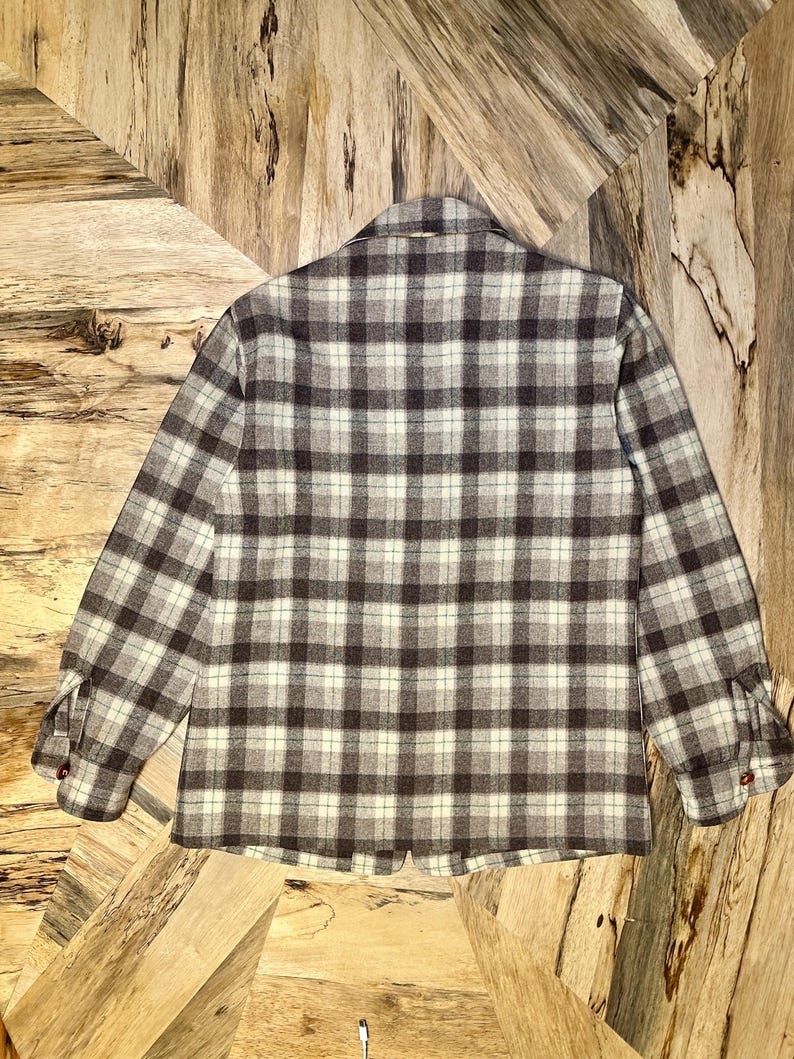 Puede incluir: Una camisa de manga larga con botones, con un dise&ntilde;o de cuadros en tonos marr&oacute;n, gris y crema. La camisa tiene cuello y pu&ntilde;os abotonados. La tela parece ser un material suave y tejido.