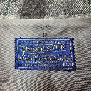 Puede incluir: Primer plano de una etiqueta Pendleton azul y dorada cosida sobre una tela gris. La etiqueta dice "PENDLETON WOOLEN MILLS, PORTLAND, OREGON, 100% VIRGIN WOOL." La tela tiene un patr&oacute;n de cuadros.