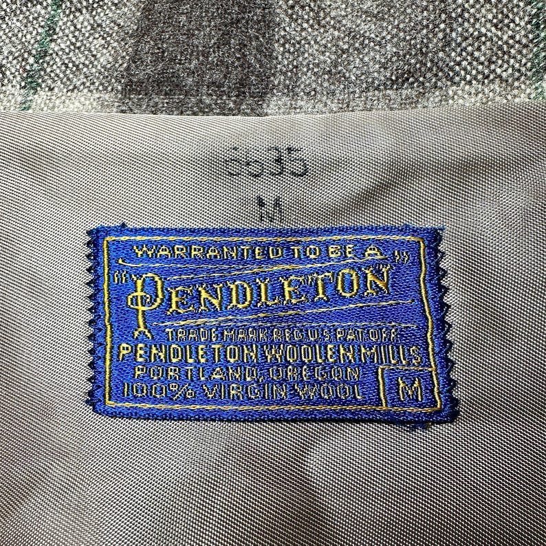 Puede incluir: Primer plano de una etiqueta Pendleton azul y dorada cosida sobre una tela gris. La etiqueta dice "PENDLETON WOOLEN MILLS, PORTLAND, OREGON, 100% VIRGIN WOOL." La tela tiene un patr&oacute;n de cuadros.