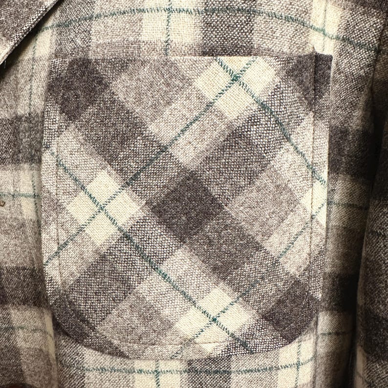 Puede incluir: Primer plano de una chaqueta a cuadros gris, marr&oacute;n y crema con un bolsillo. La chaqueta tiene un patr&oacute;n de cuadros cl&aacute;sico con una mezcla de colores y texturas. El bolsillo es de un tono marr&oacute;n m&aacute;s oscuro y la costura es visible.