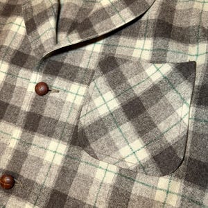 Puede incluir: Primer plano de una chaqueta a cuadros en tonos marr&oacute;n, gris y crema, con sutiles acentos verdes. La chaqueta presenta un cuello con solapas, un bolsillo en el pecho y dos botones de cuero marr&oacute;n. La tela tiene un aspecto texturizado.