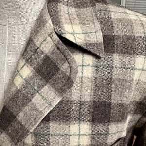 Puede incluir: Primer plano de una blazer de tweed con estampado de cuadros. La chaqueta presenta una mezcla de cuadrados grises, crema y marr&oacute;n oscuro con finas l&iacute;neas turquesas. La solapa de la blazer es visible, mostrando el dise&ntilde;o a medida.