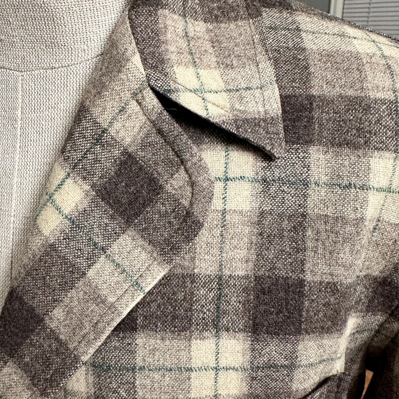 Puede incluir: Primer plano de una blazer de tweed con estampado de cuadros. La chaqueta presenta una mezcla de cuadrados grises, crema y marr&oacute;n oscuro con finas l&iacute;neas turquesas. La solapa de la blazer es visible, mostrando el dise&ntilde;o a medida.