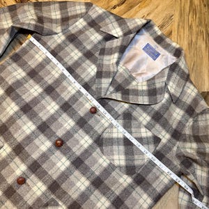 Puede incluir: Chaqueta vintage a cuadros en tonos grises, crema y marr&oacute;n. La chaqueta presenta un patr&oacute;n de cuadros cl&aacute;sico, un cuello y tres botones marrones. Una cinta m&eacute;trica se coloca sobre la chaqueta, mostrando medidas en cent&iacute;metros.