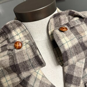 Puede incluir: Primer plano de una chaqueta a cuadros con botones de cuero marr&oacute;n. La chaqueta presenta una paleta de colores neutros de gris, beige y un toque de verde. Los botones son redondos y tienen un dise&ntilde;o de puntada cruzada.