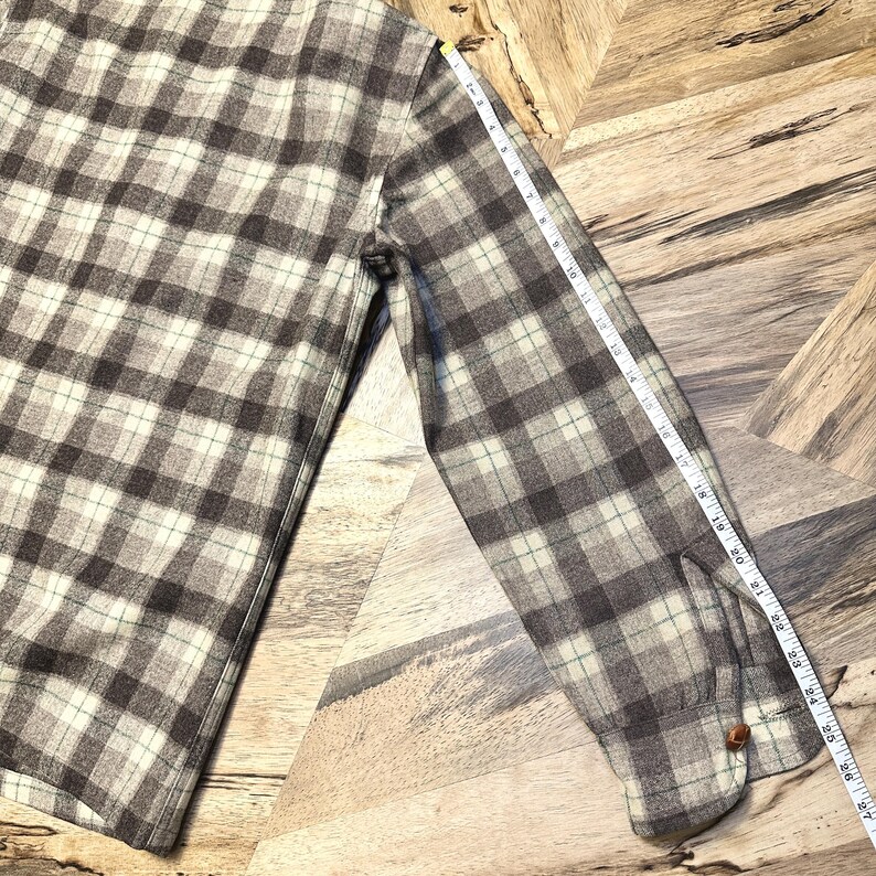 Puede incluir: Una camisa de franela abotonada de manga larga con un estampado de cuadros marr&oacute;n, crema y azul claro. La camisa se mide con una cinta m&eacute;trica, mostrando una longitud de aproximadamente 69 cm desde el hombro hasta el pu&ntilde;o.
